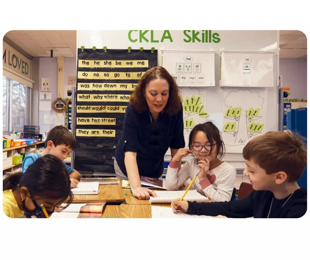 Teacher assisting four young students with reading tasks at a table in a classroom with educational posters on the wall.