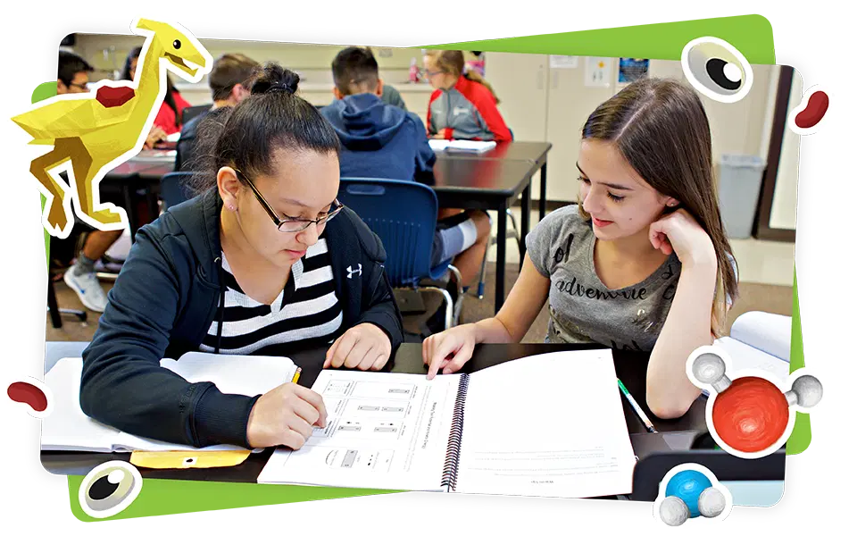 Two girls sit at a classroom desk, working together on worksheets. Other students are visible in the background. Cartoon science-themed graphics decorate the image border.